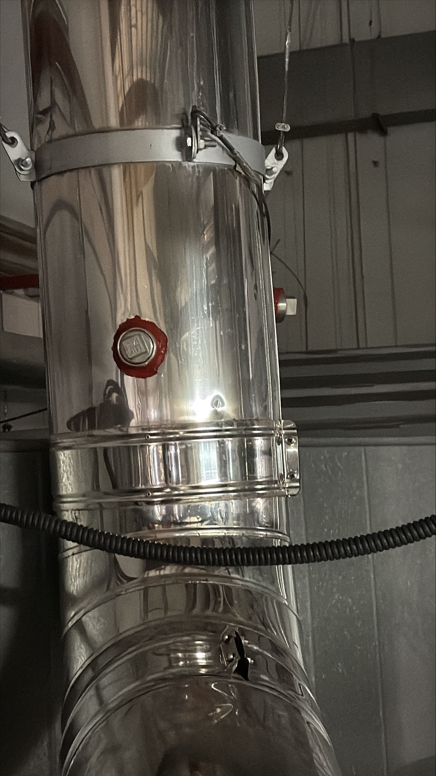 TYPE: photograph
CONTENT: The image shows a close-up of a stainless steel pipe, likely an exhaust or ventilation duct, in an industrial or utility setting. It