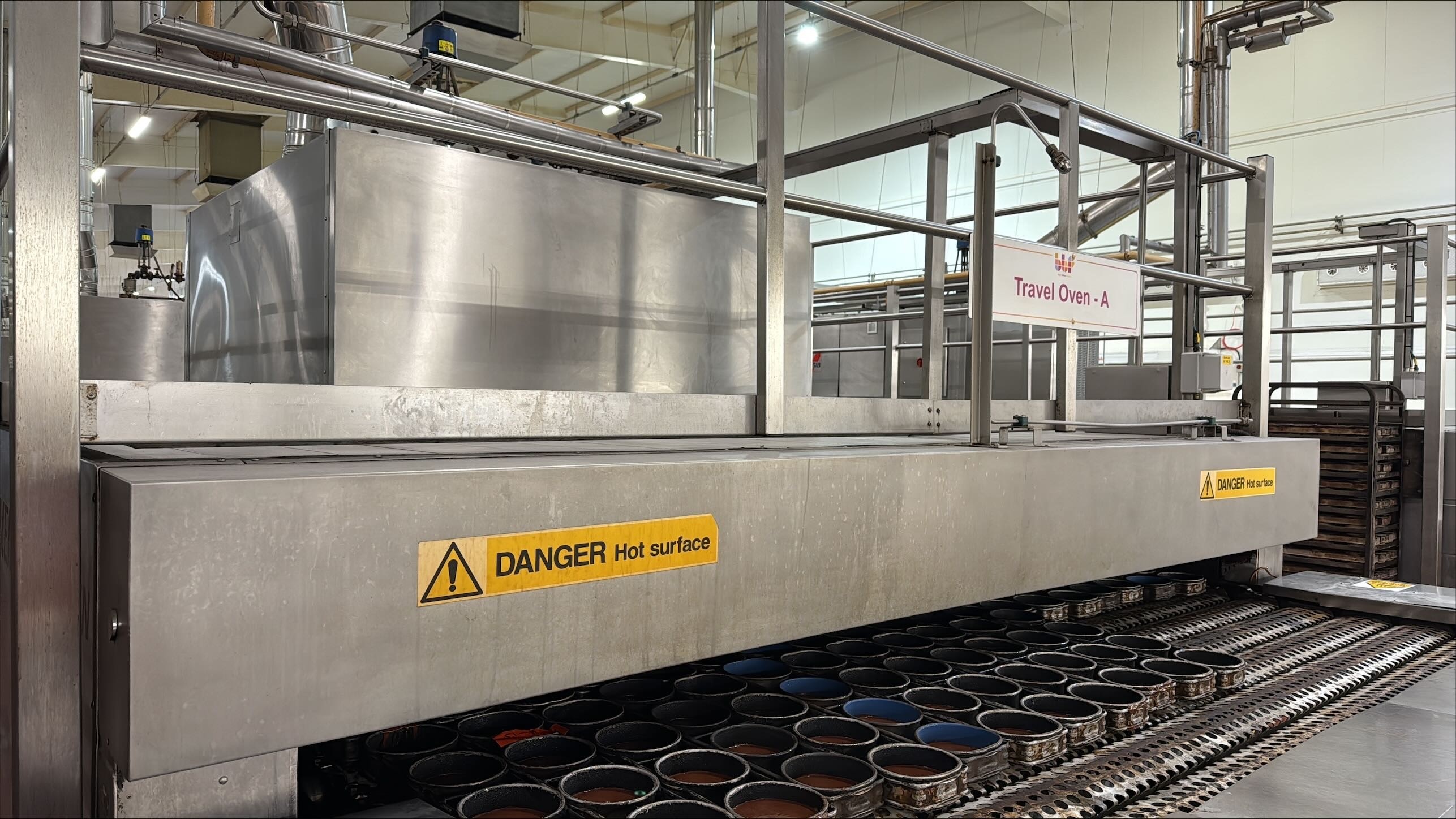 TYPE: photograph
CONTENT: This image displays an industrial factory setting, specifically a commercial bakery, featuring a large "Travel Oven - A