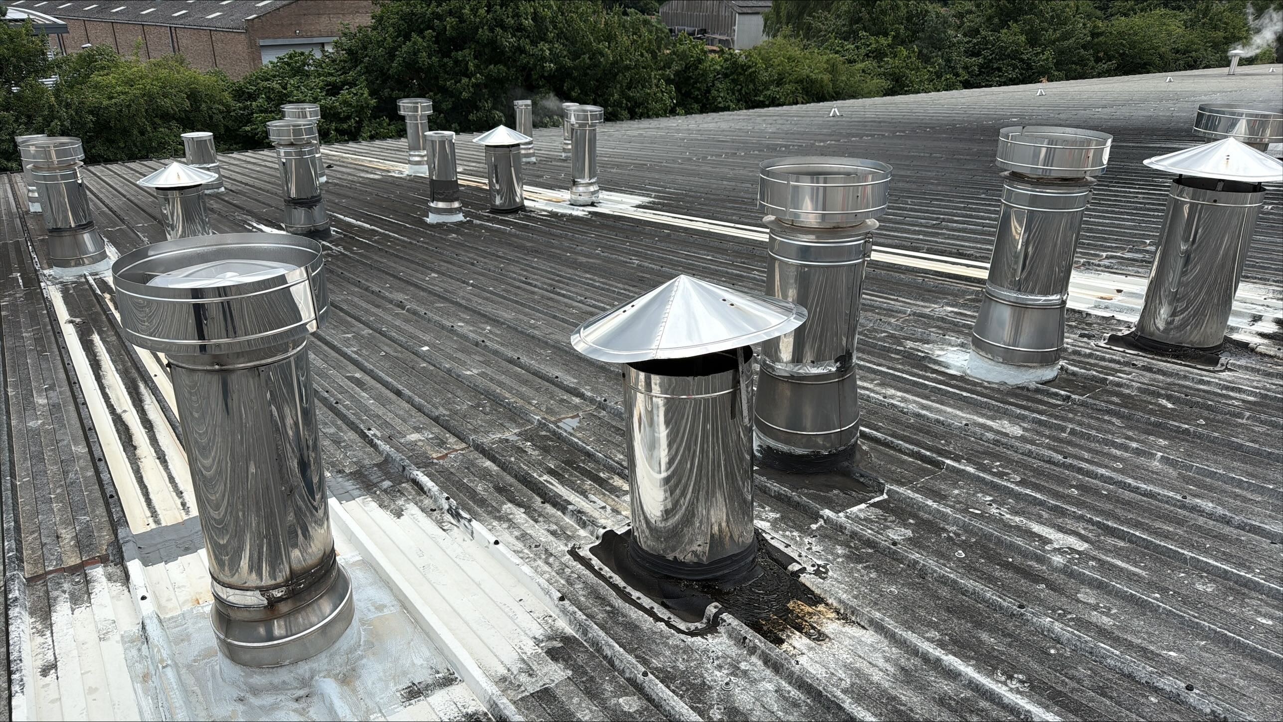 TYPE: photograph
CONTENT: The image shows an aerial view of multiple stainless steel chimneys/vents installed on a corrugated metal roof, with various sealants and some signs of wear or leakage around their bases. In the background, trees and industrial buildings are visible under a bright sky.
QUALITY: high
