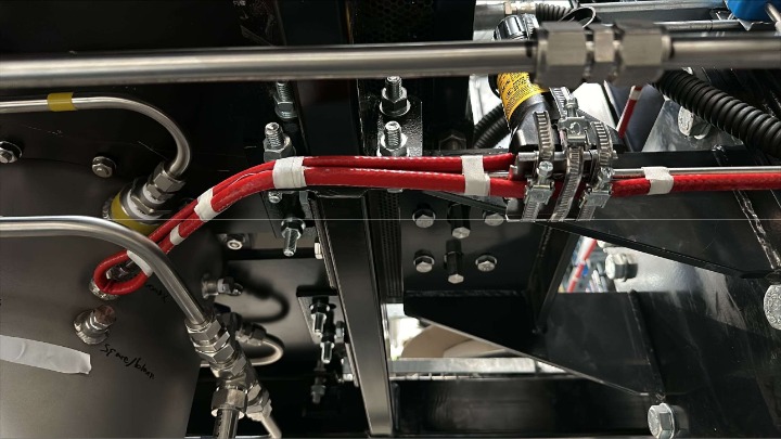 TYPE: photograph
CONTENT: This photograph shows a close-up view of industrial or scientific equipment, featuring metallic pipes/tubing, red cables secured with white tape and metal clamps, and various bolts and nuts on