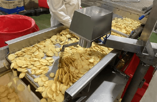 A product bed of ridged potato slices travels on stainless-steel conveyors through continuous frying equipment. The image shows the product flow whose line speed is adjusted in the oil content control loop, affecting time spent in the oil.